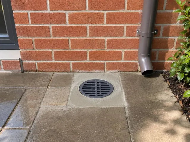 Outdoor drain grate set in paved area beside a brick wall and downpipe in Rhodes